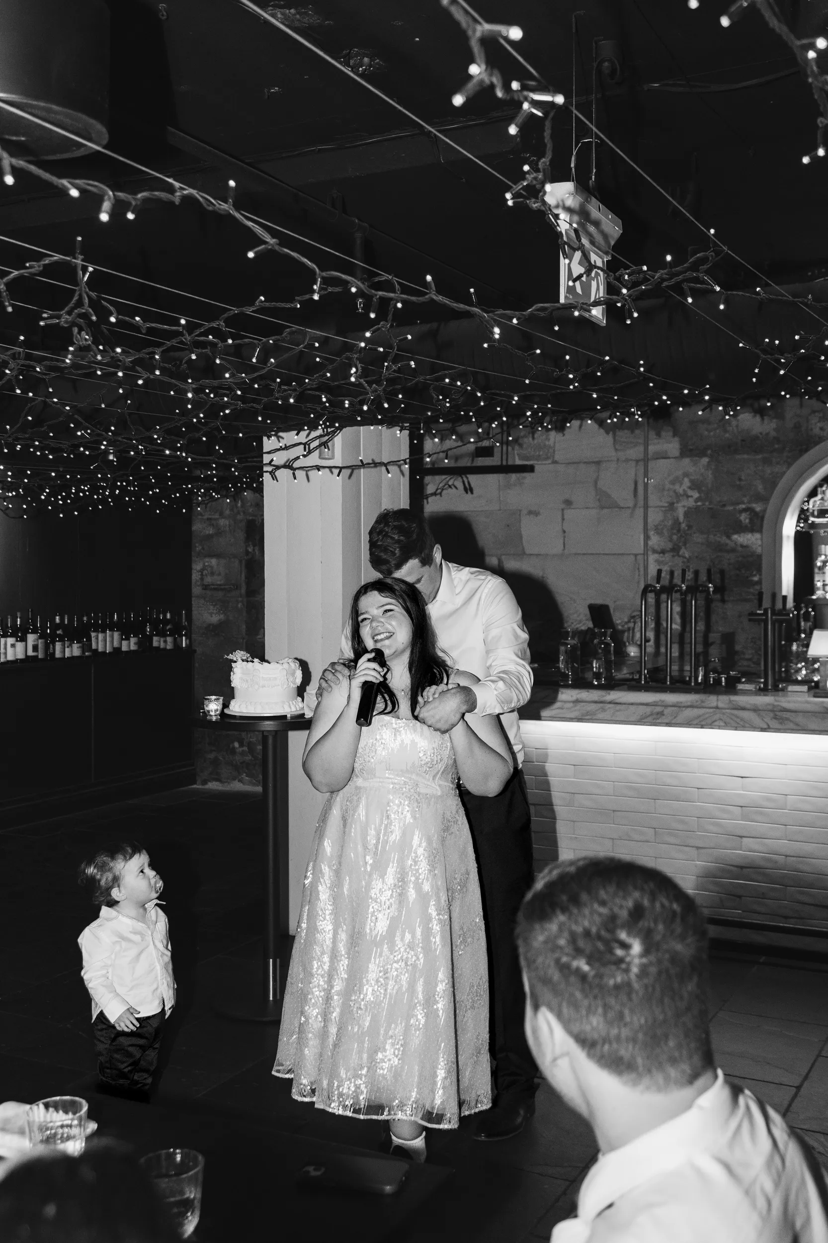 Carlie giving heartfelt speech with microphone in rustic cellar venue with string lights while Iain supports her