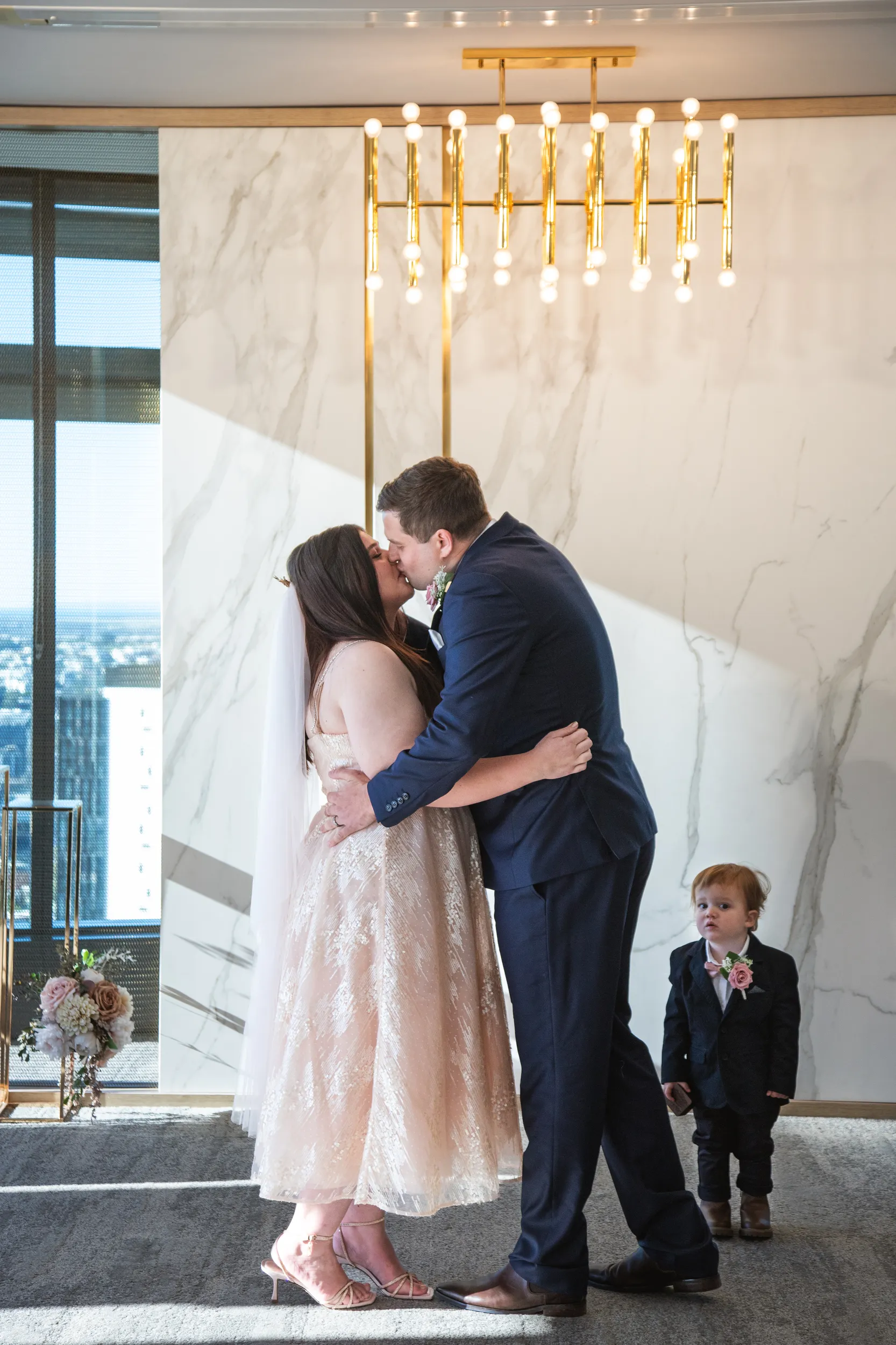 Carlie and Iain heartwarming ceremony kiss under elegant gold chandeliers with young child watching