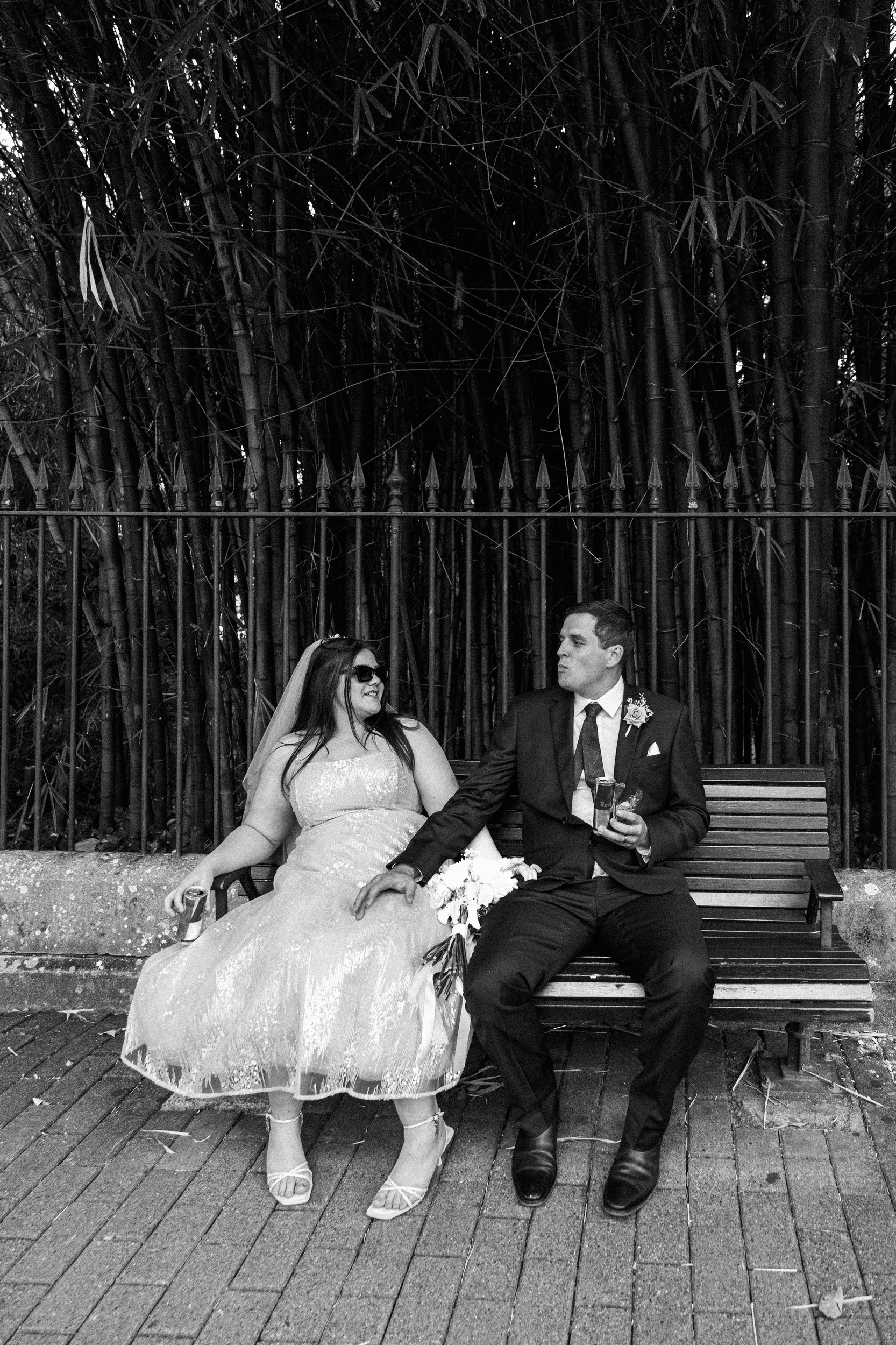 Carlie and Iain relaxed black and white moment sitting on wooden bench with drinks and sunglasses