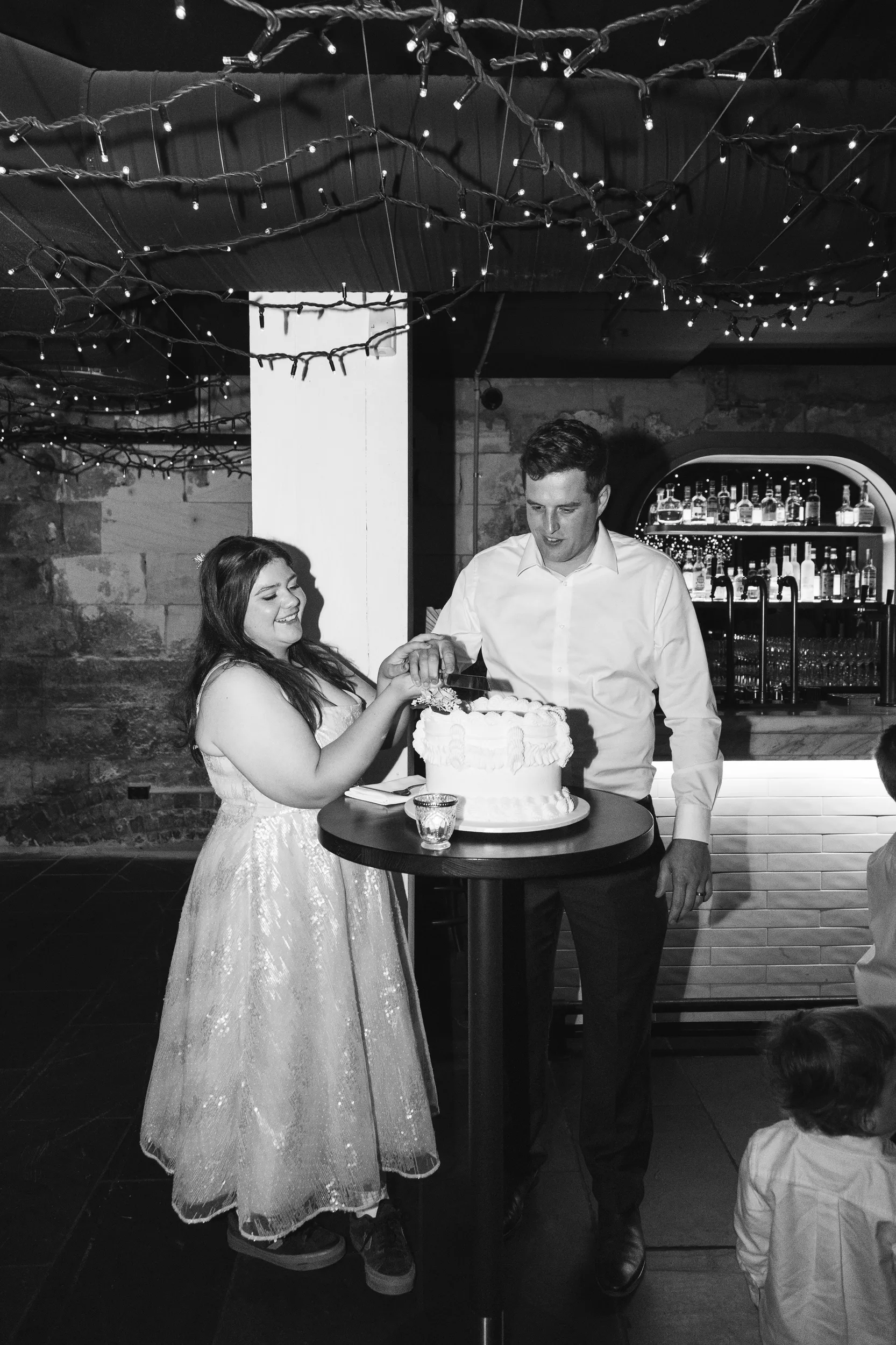 Carlie and Iain black and white cake cutting ceremony in rustic cellar venue with string lights and exposed brick