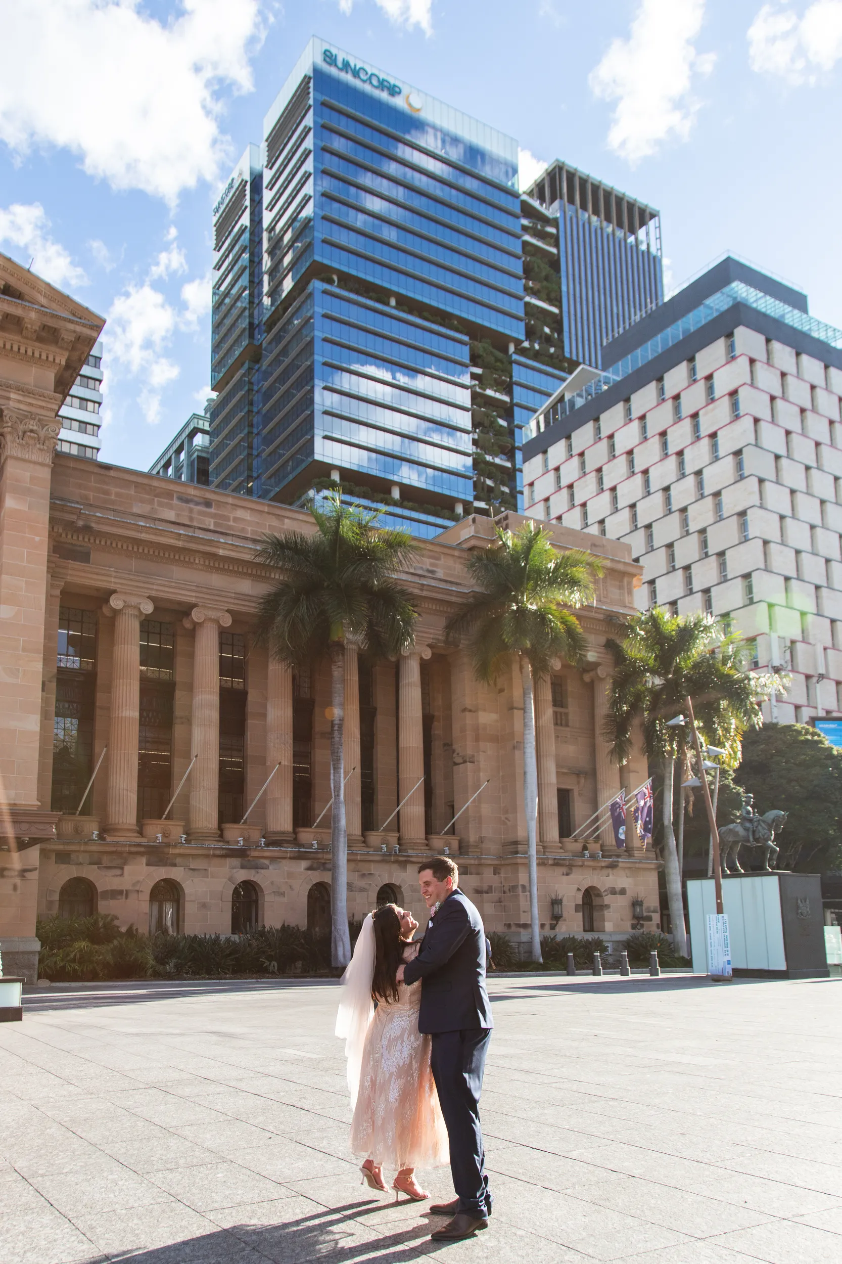 Carlie and Iain romantic embrace in front of grand classical building with modern urban skyline backdrop