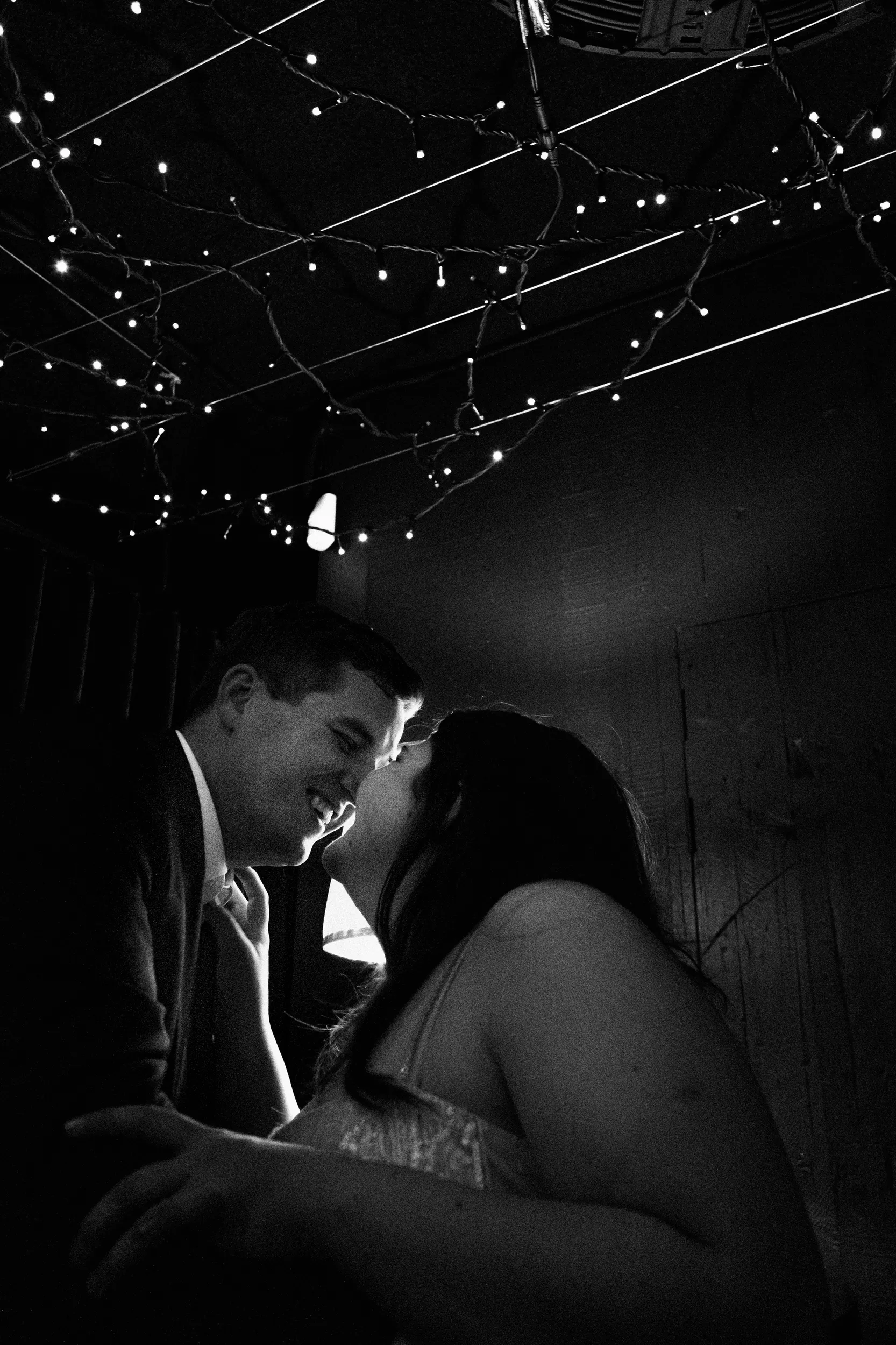 Carlie and Iain intimate black and white moment about to kiss in rustic cellar with dramatic string lights overhead