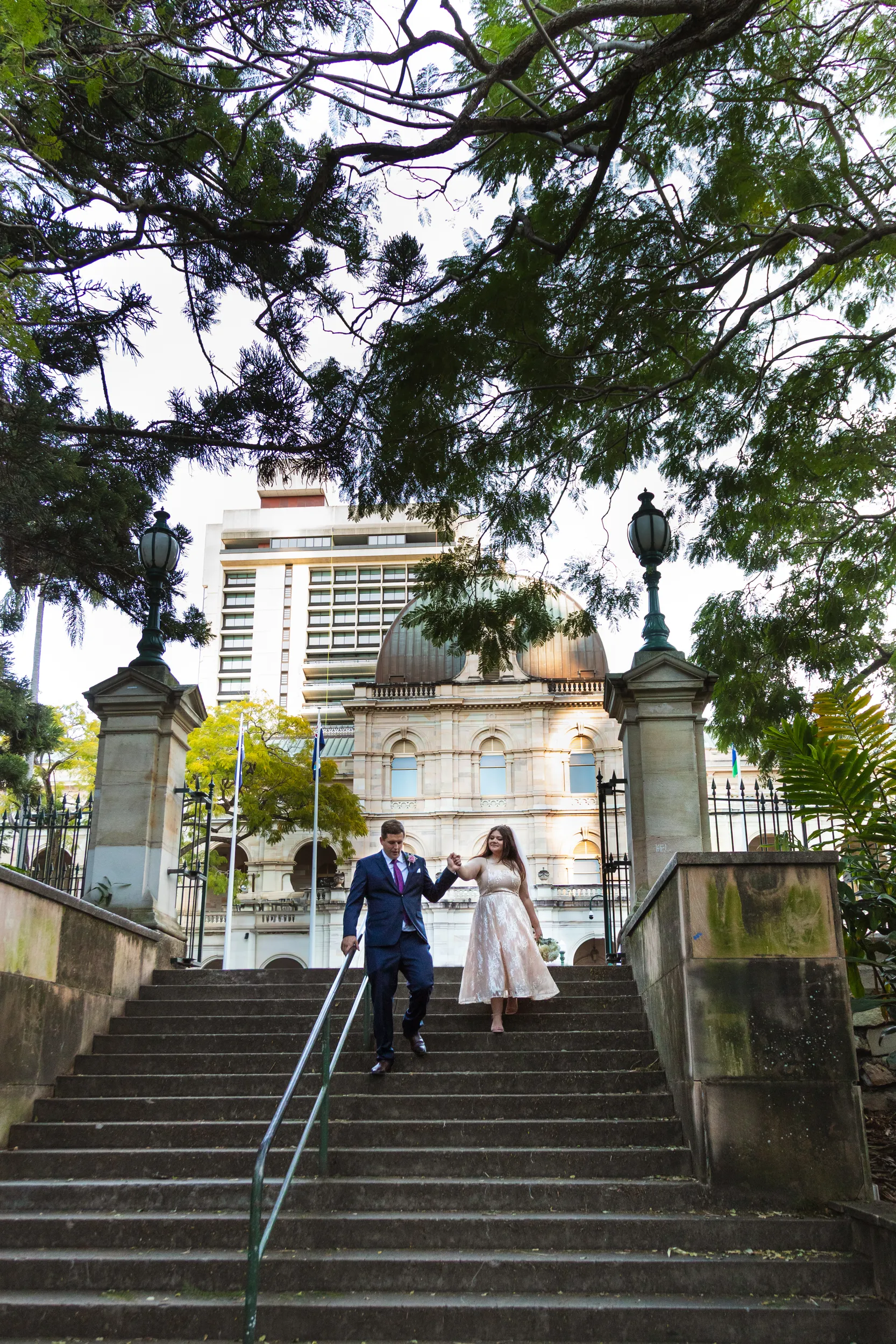 Carlie and Iain walking hand-in-hand down stone steps in front of historic building with city skyline