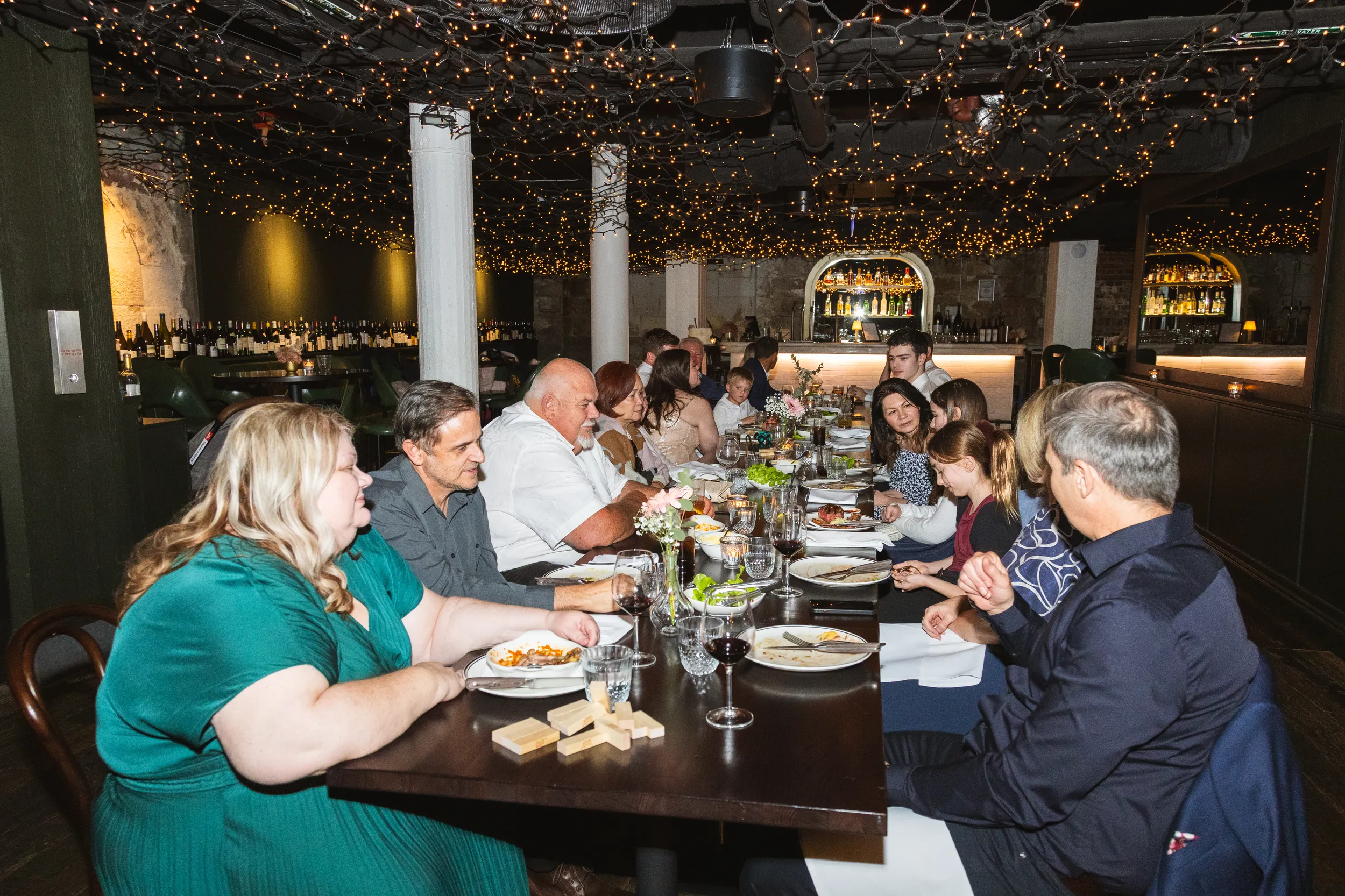 Wedding reception dinner with extended family and friends gathered around long table in rustic cellar with string lights