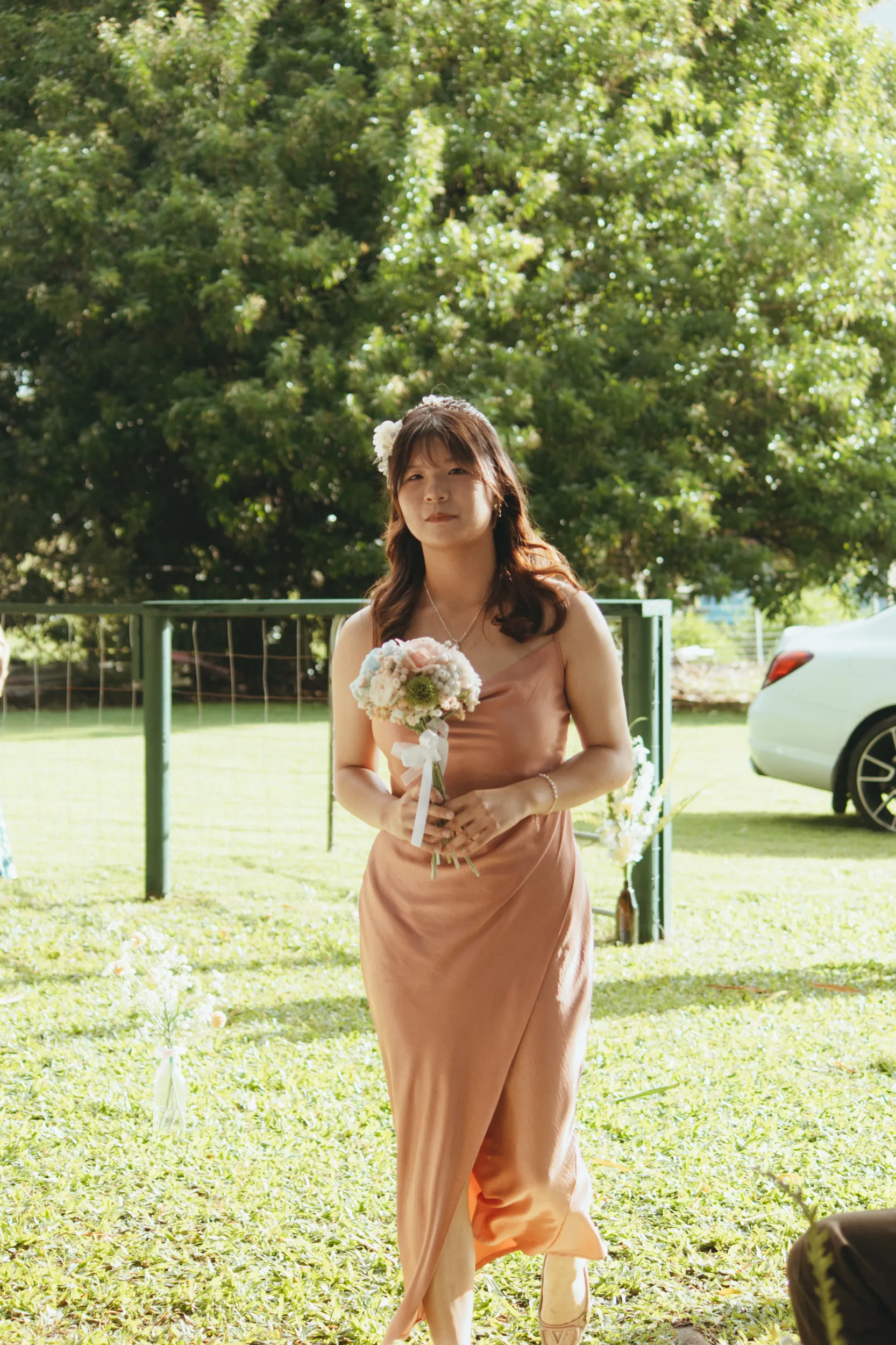 Bridesmaid in champagne colored dress walking with small bouquet, wearing delicate headpiece and pearl necklace