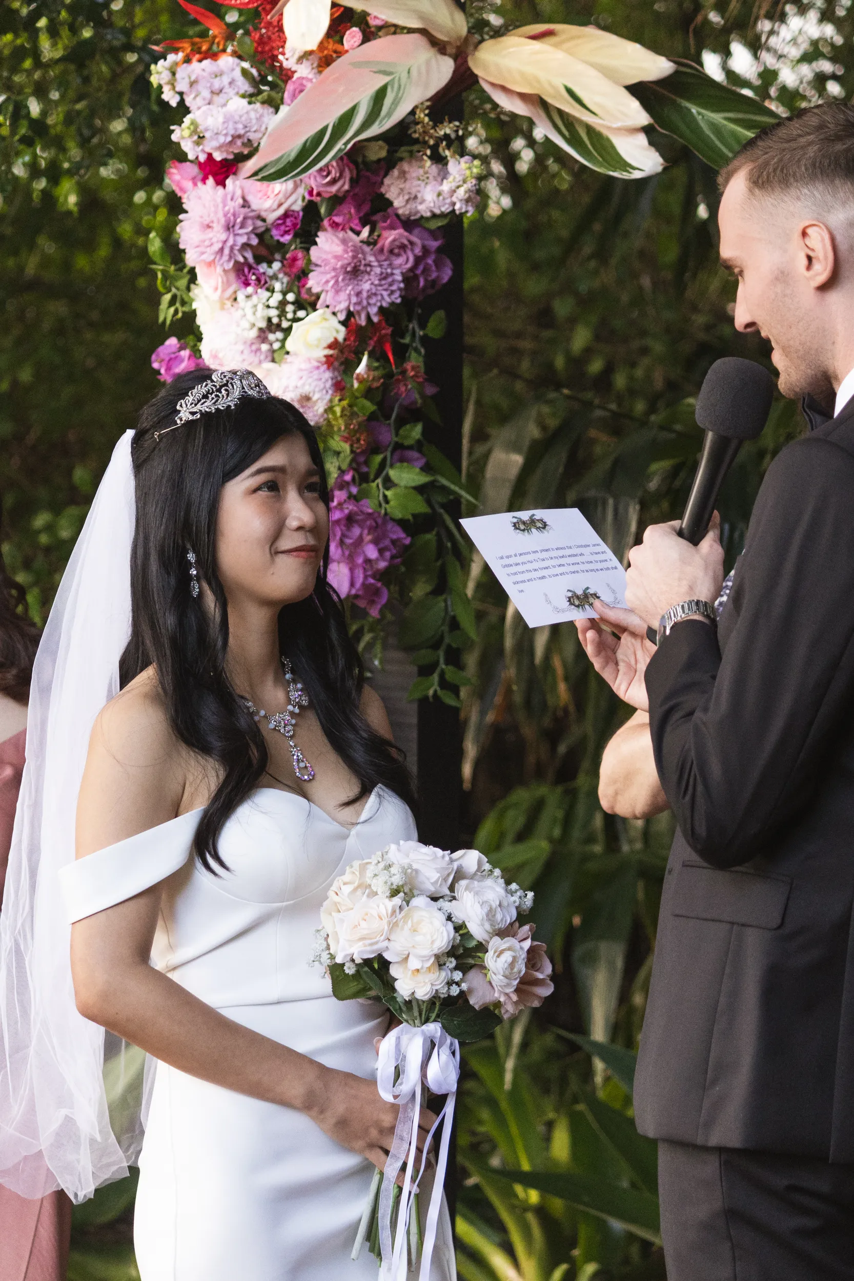 Chris reading wedding vows into microphone during ceremony with Huiyu listening surrounded by vibrant purple and pink floral arrangements