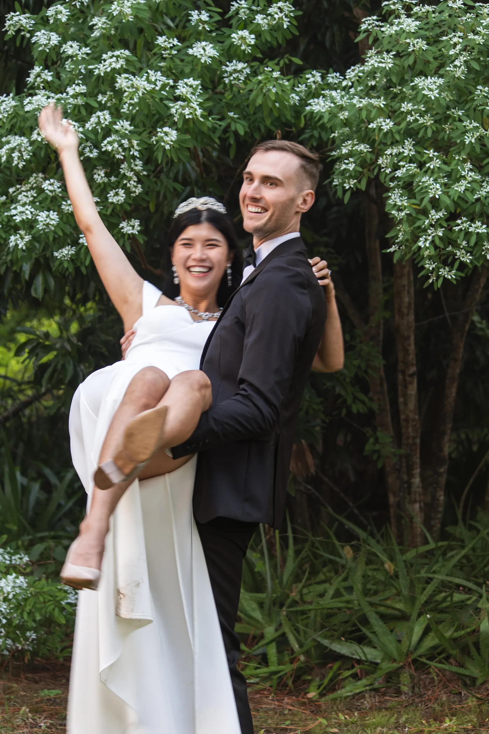Huiyu and Chris joyful moment with groom carrying bride while she raises hand in celebration against white flowering trees