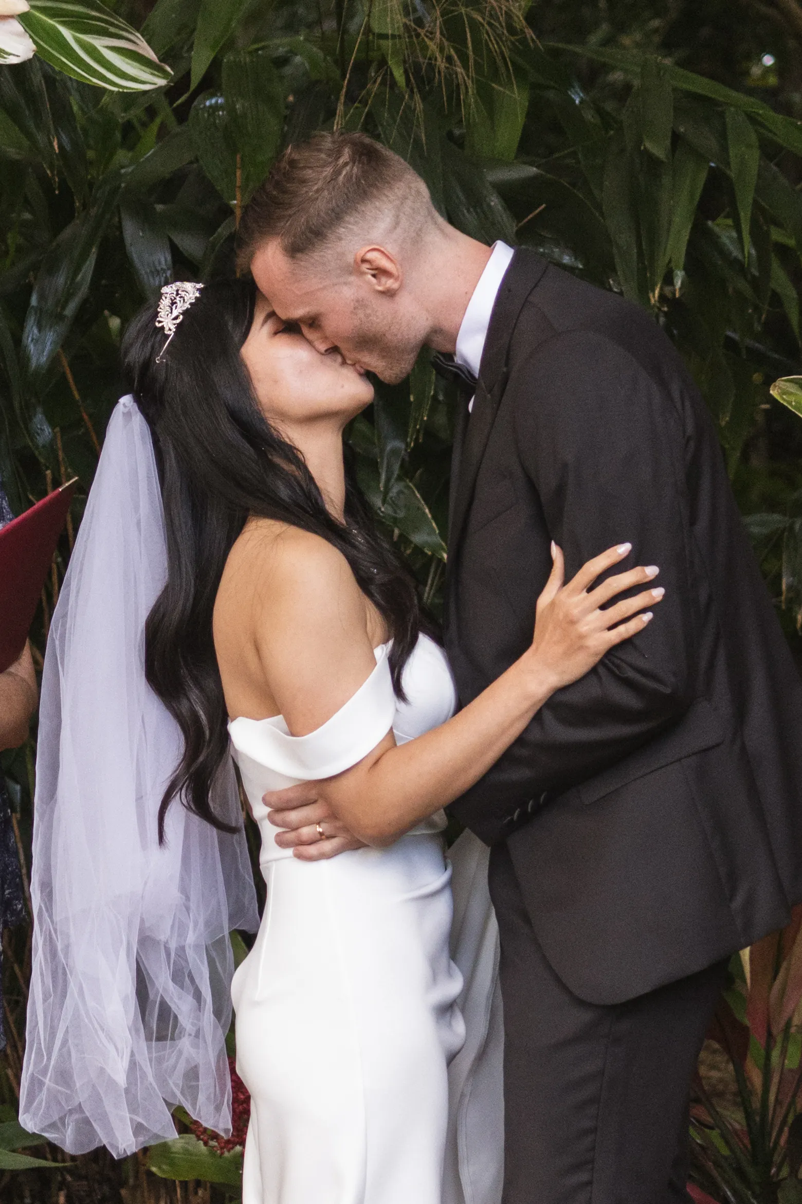 Huiyu and Chris romantic garden kiss with bride in white off-shoulder dress and crystal tiara surrounded by lush tropical foliage