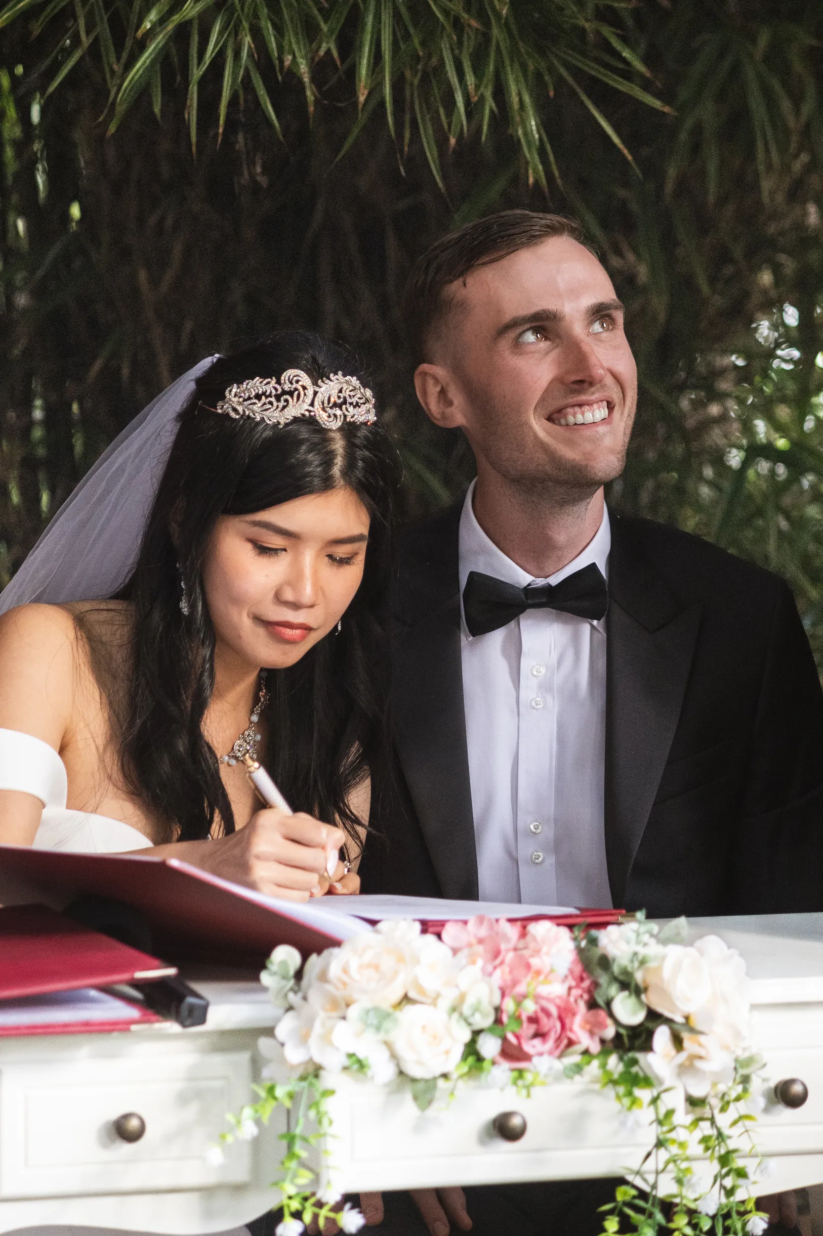 Huiyu and Chris signing marriage certificate with bride in crystal tiara and groom smiling joyfully
