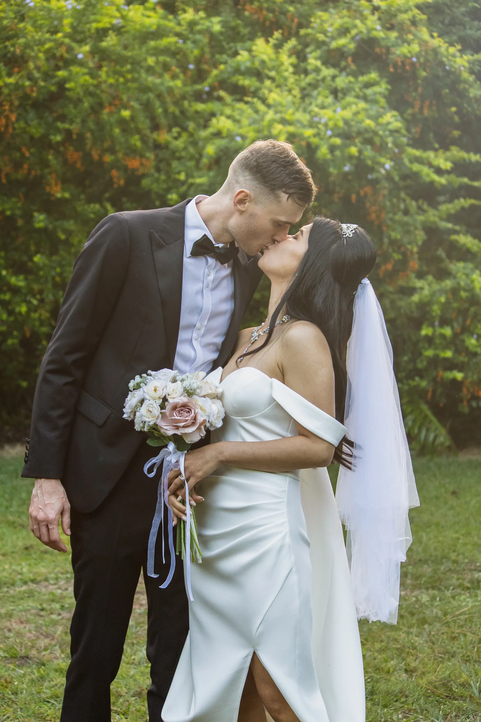 Huiyu and Chris tender kiss in garden with bride holding white rose and dusty pink bouquet in intimate embrace