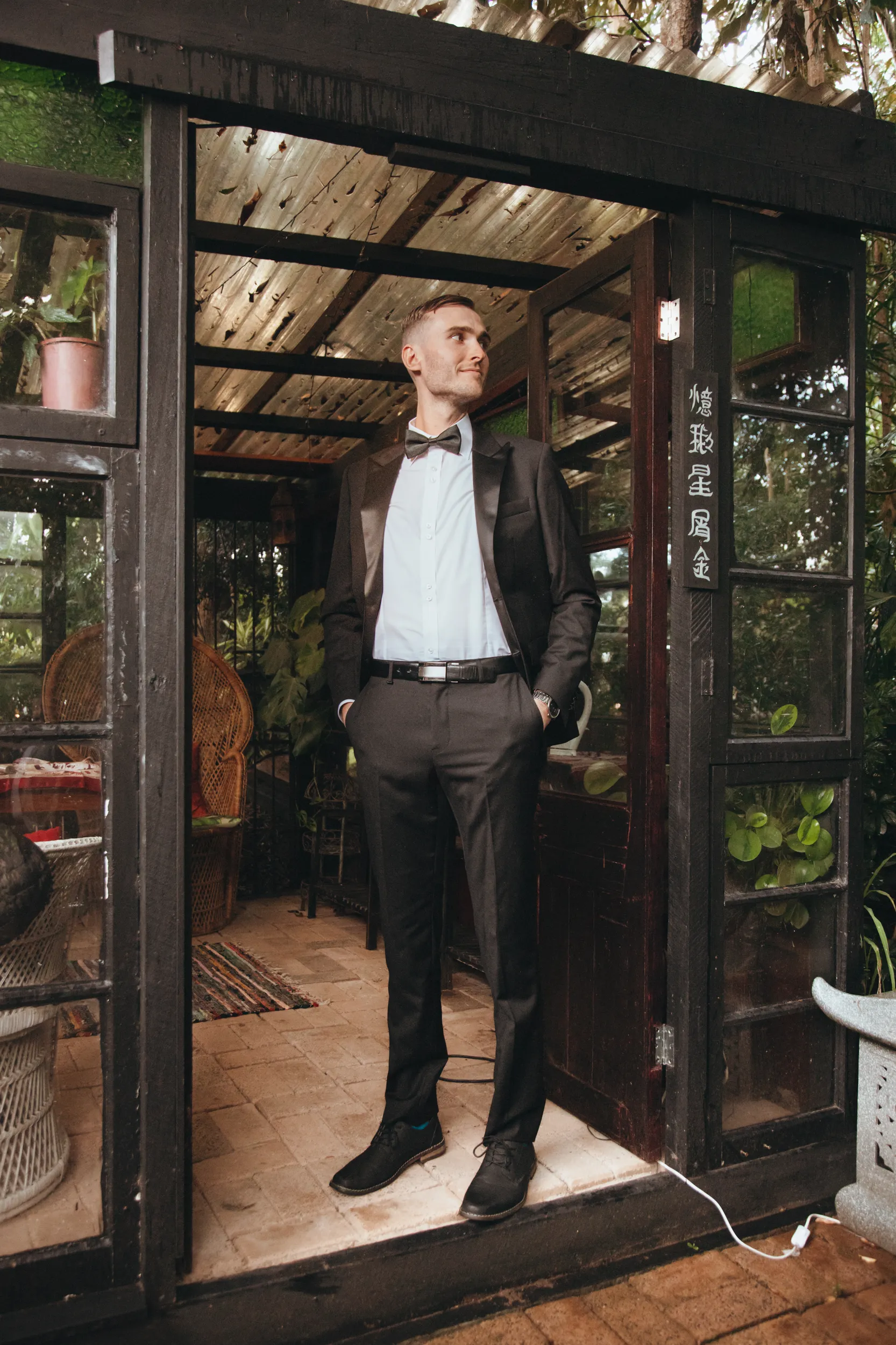 Chris stylish groom portrait in dark suit and bow tie in front of rustic building with Chinese characters on window