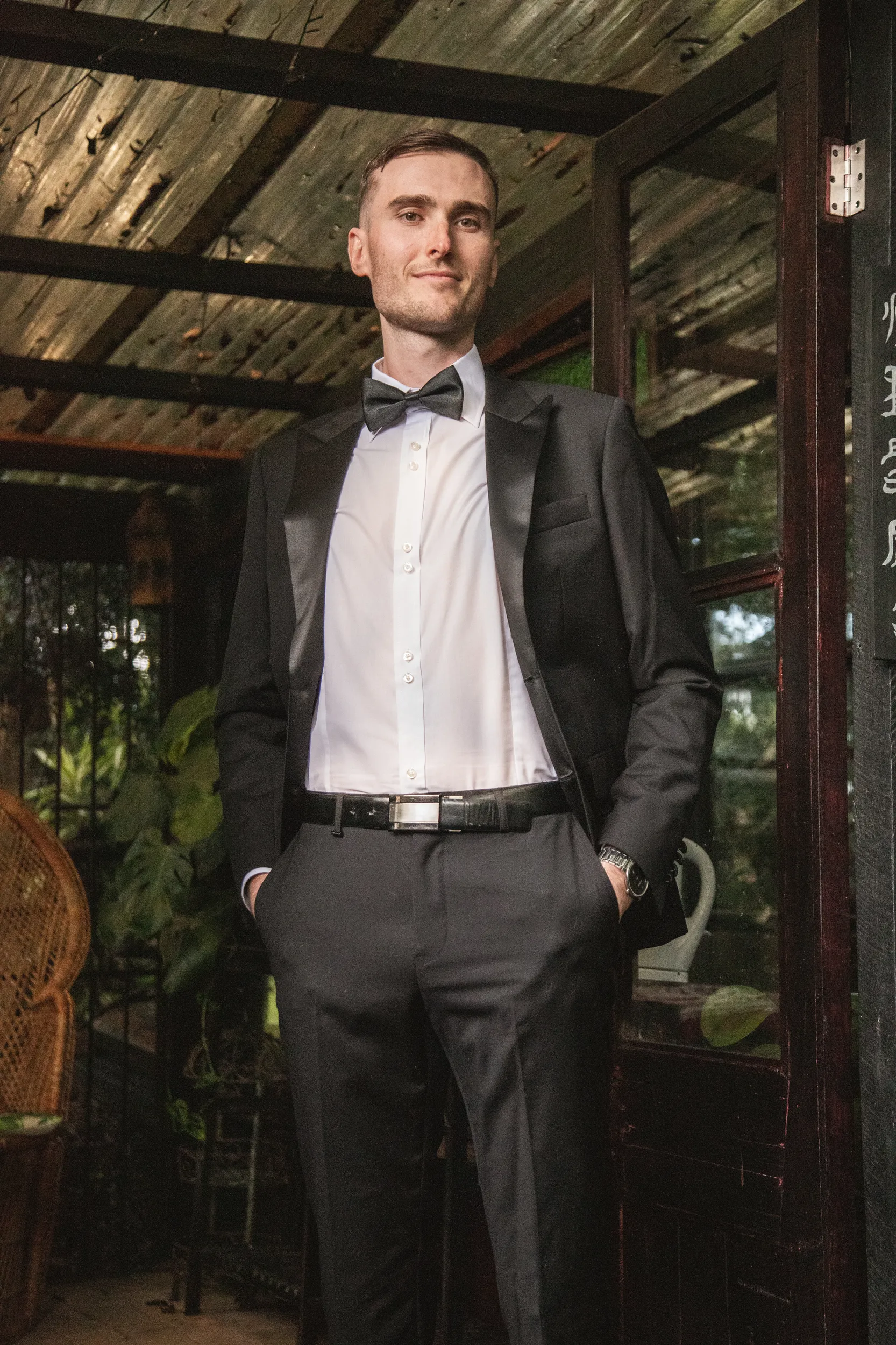 Chris stylish groom portrait in black tuxedo with bow tie in rustic wooden structure with natural lighting
