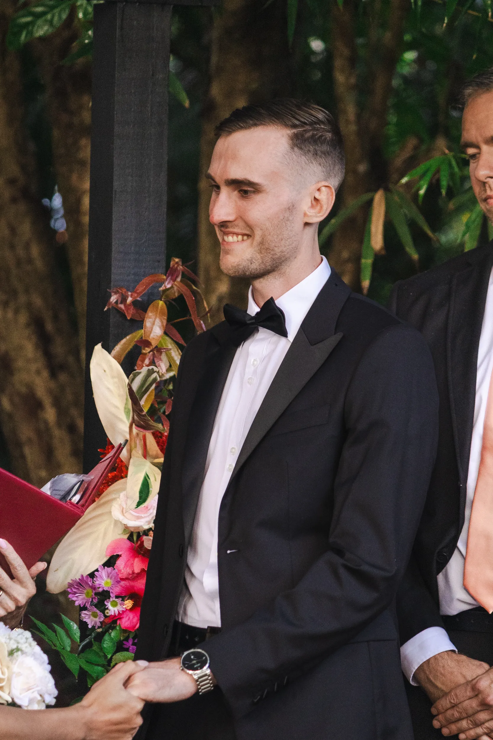 Chris candid moment receiving colorful bouquet while seated in tuxedo with natural garden lighting