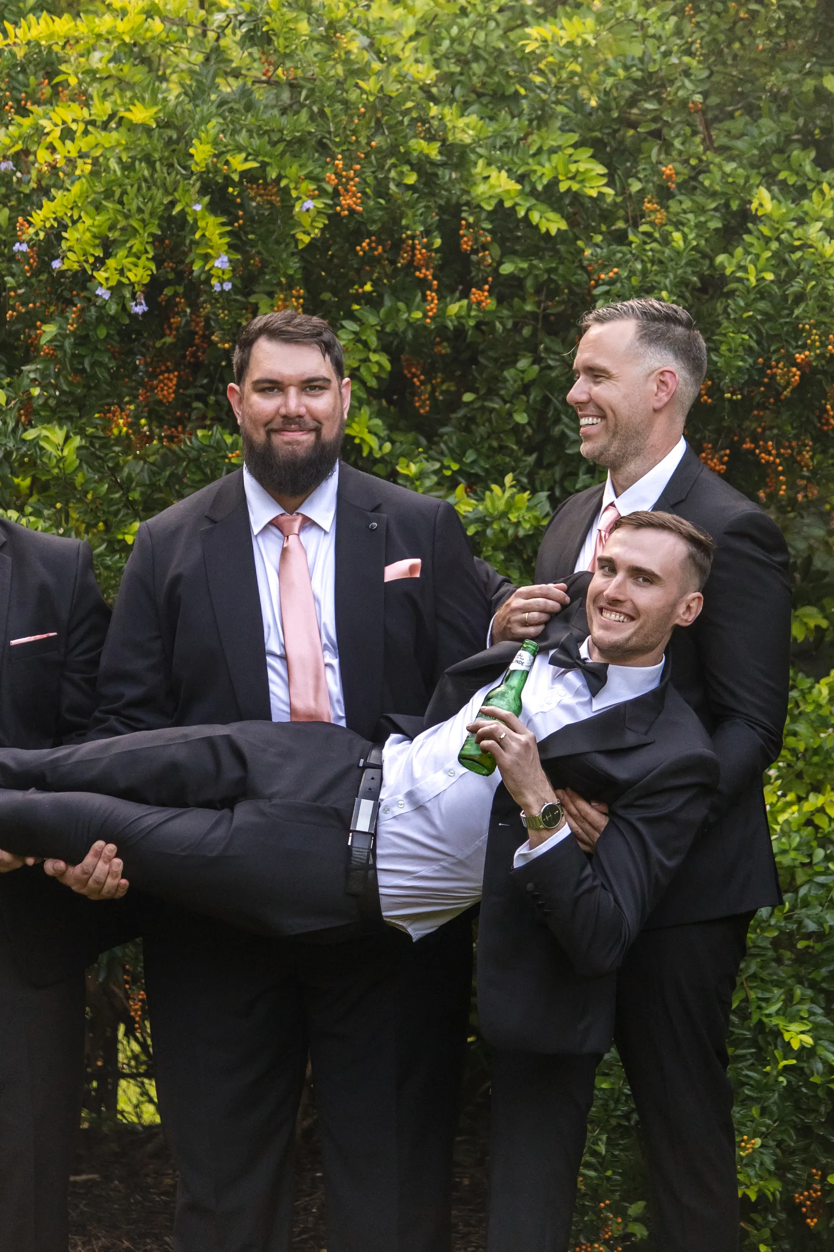 Huiyu and Chris wedding groomsmen playful moment lifting friend with beer bottle in vibrant garden