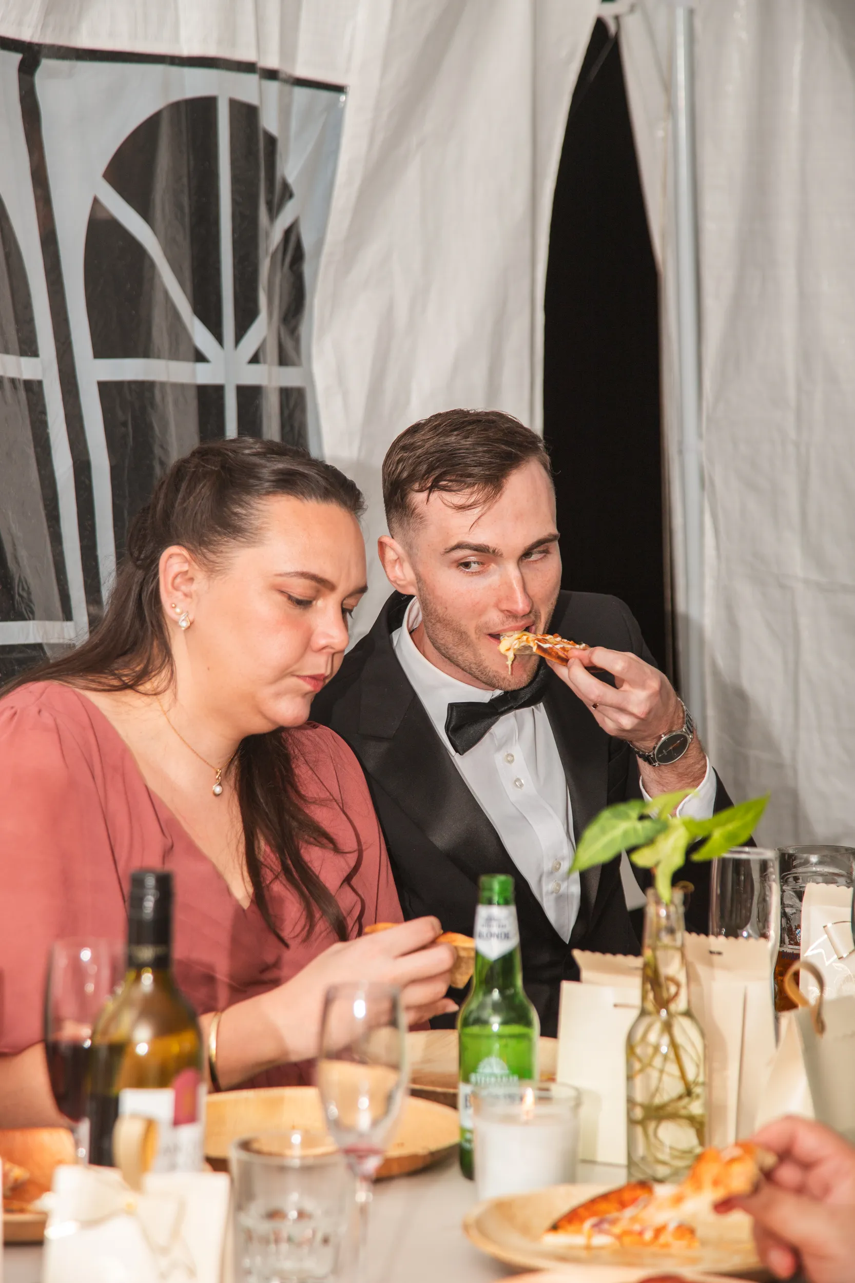 Chris candid reception moment eating pizza in tuxedo sitting next to woman in coral dress