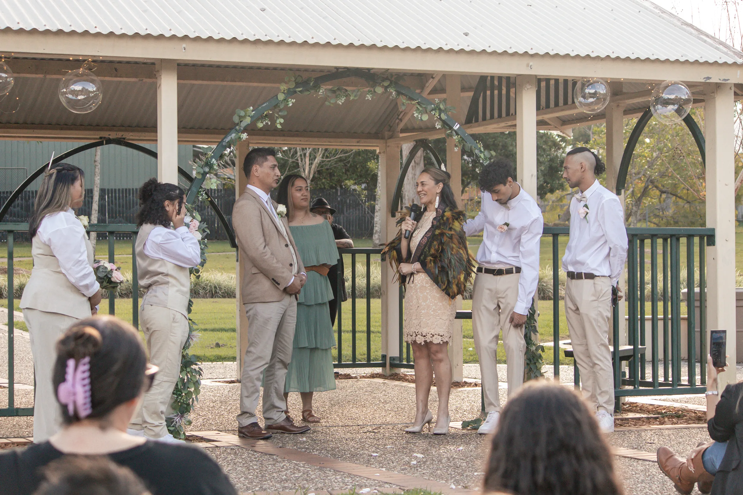 Wide ceremony shot with bride speaking into microphone surrounded by wedding party and guests