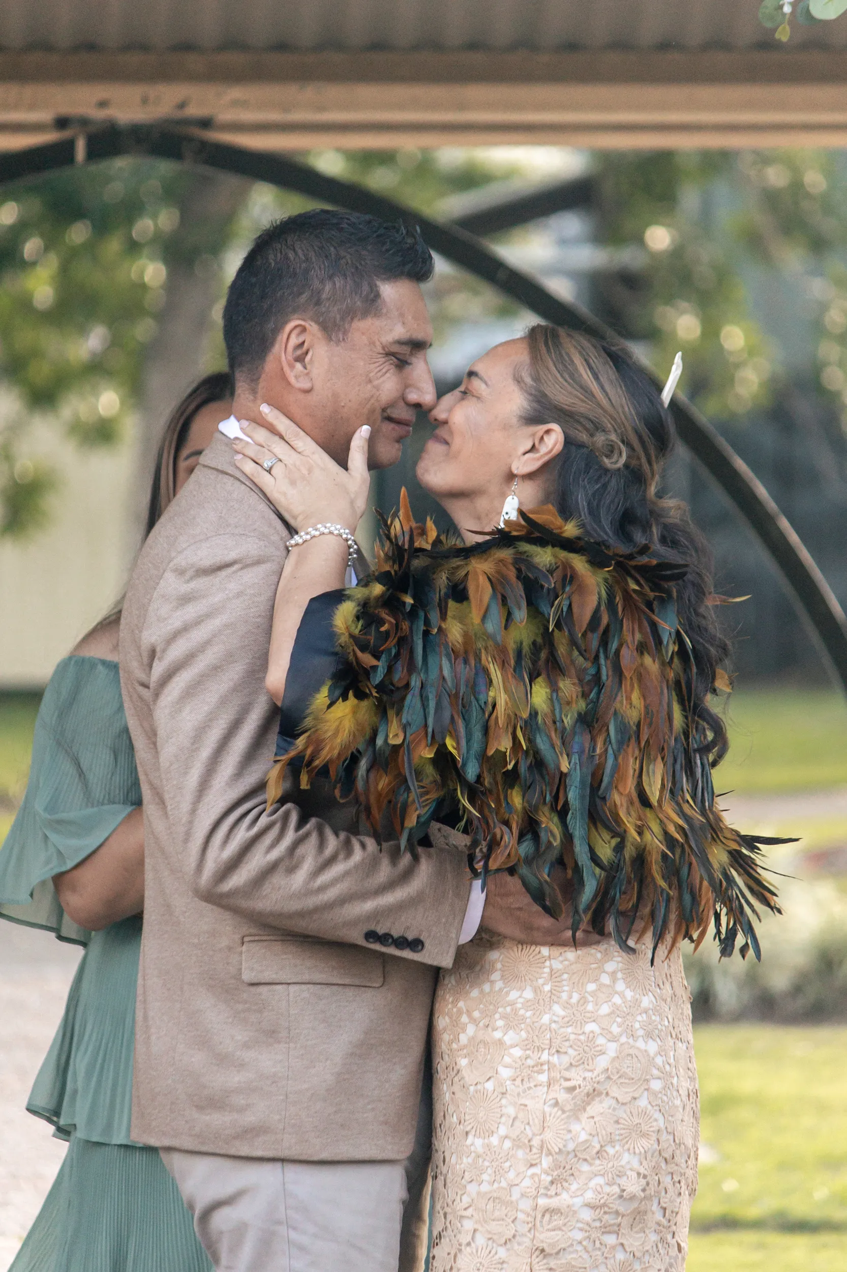 Intimate couple embrace under floral arch with bride in traditional Māori feather cloak