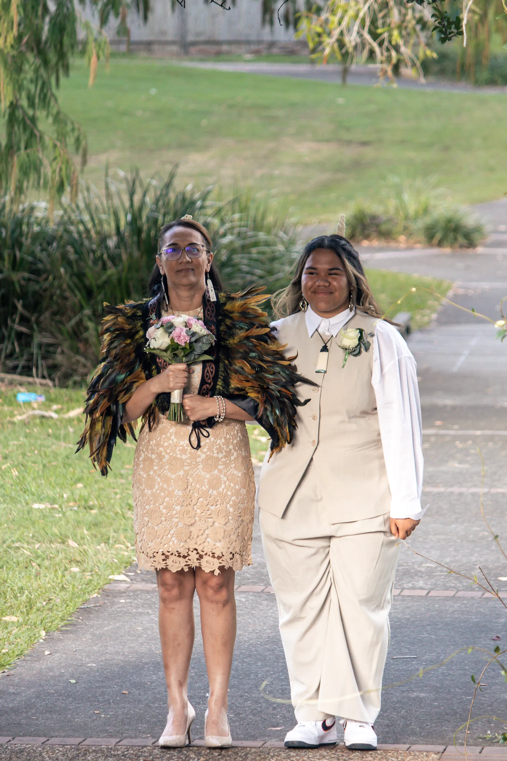 Beautiful bridal portrait in traditional Māori feather cloak holding bouquet with companion on park pathway