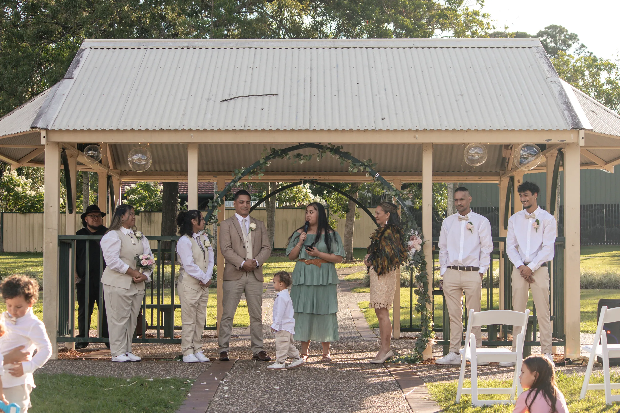 Wedding ceremony moment under pavilion with bride in traditional feather cloak and family members