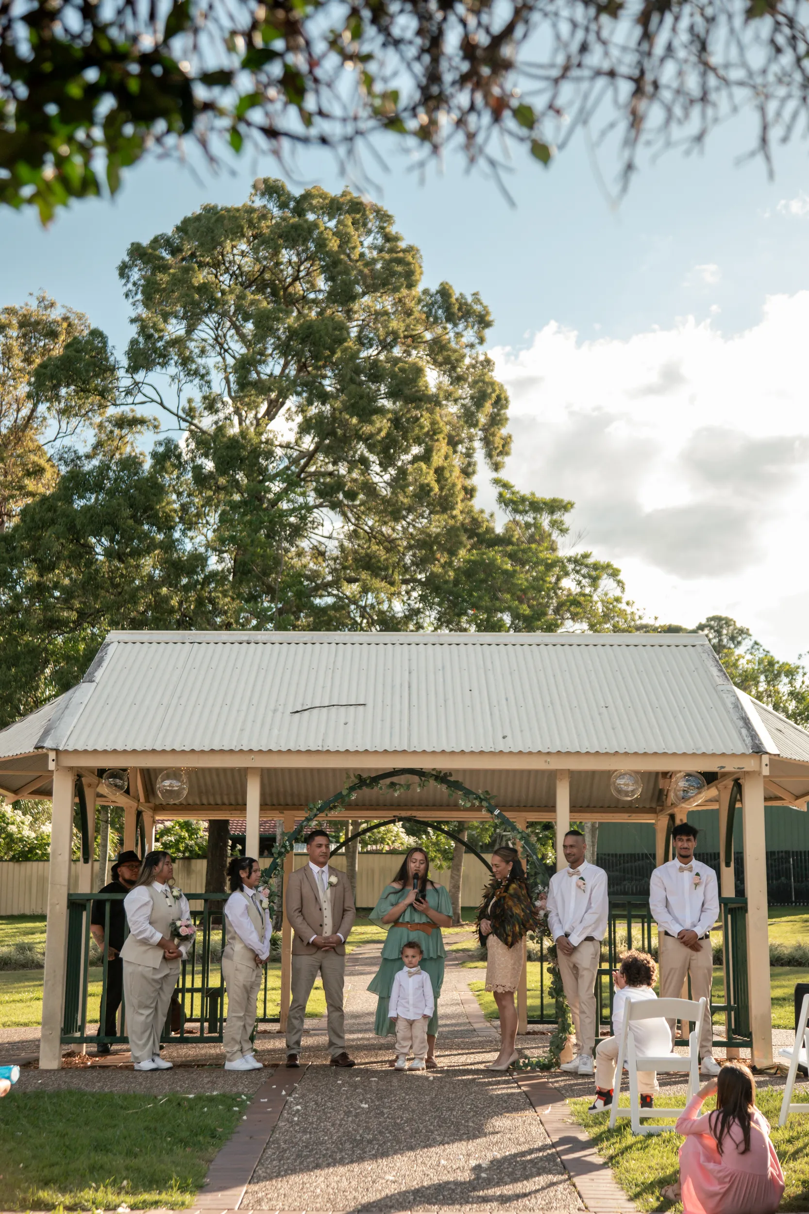 Wide shot of outdoor wedding ceremony under rustic pavilion with children playing and guests seated