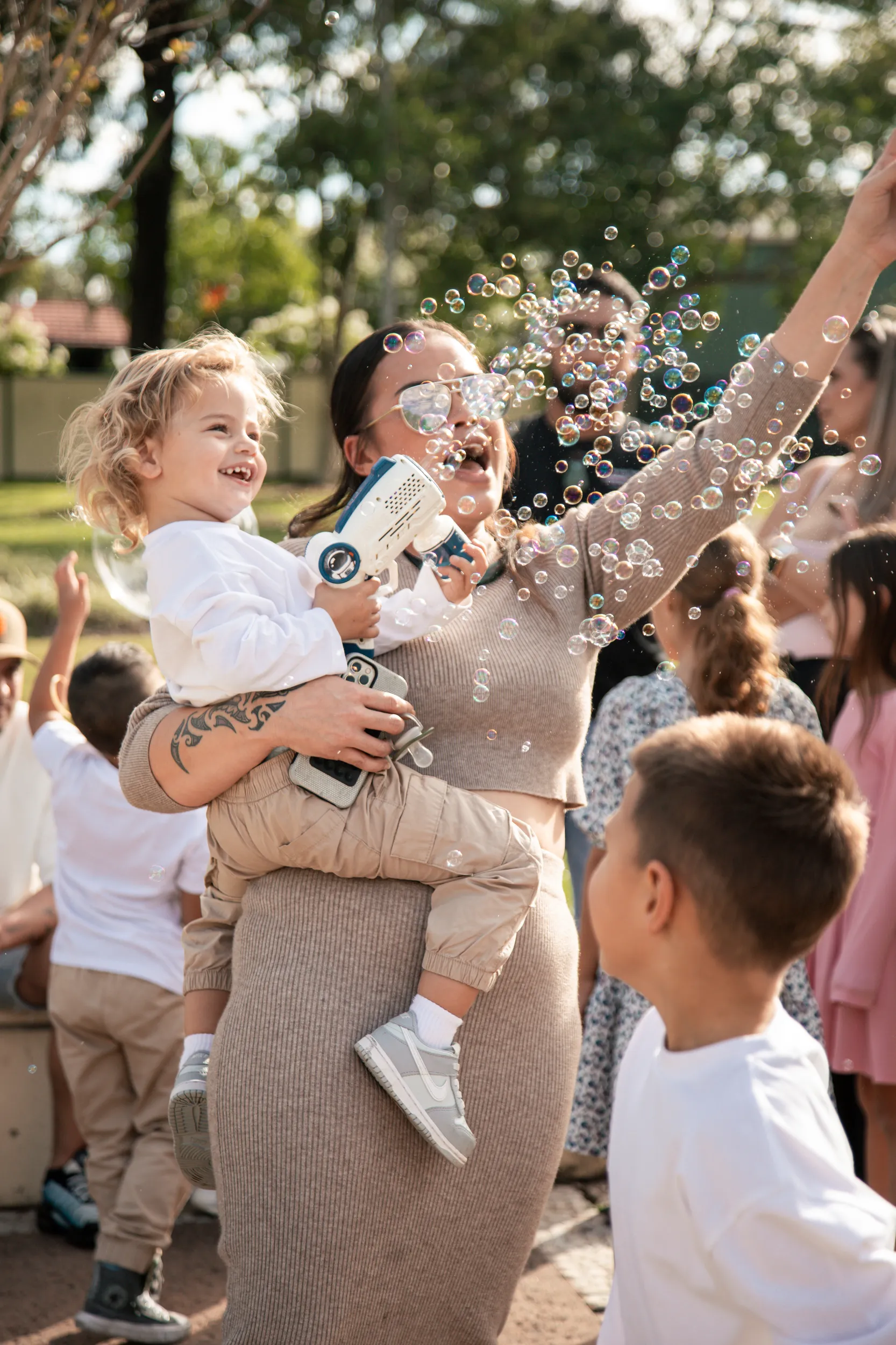 Joyful wedding celebration with family member holding child creating magical soap bubbles in park during golden hour