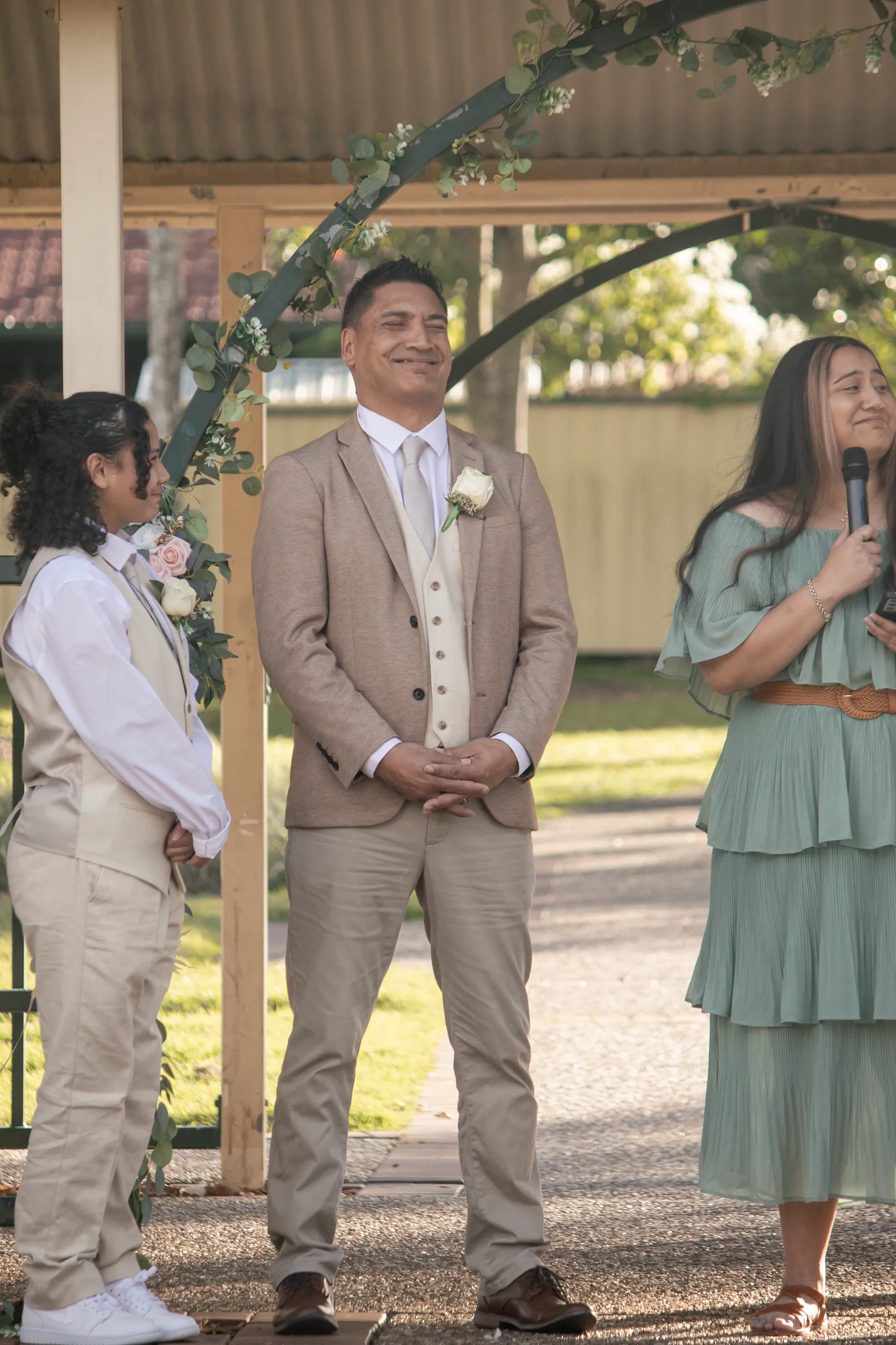 Happy groom smiling during wedding ceremony under floral arch with officiant and wedding party
