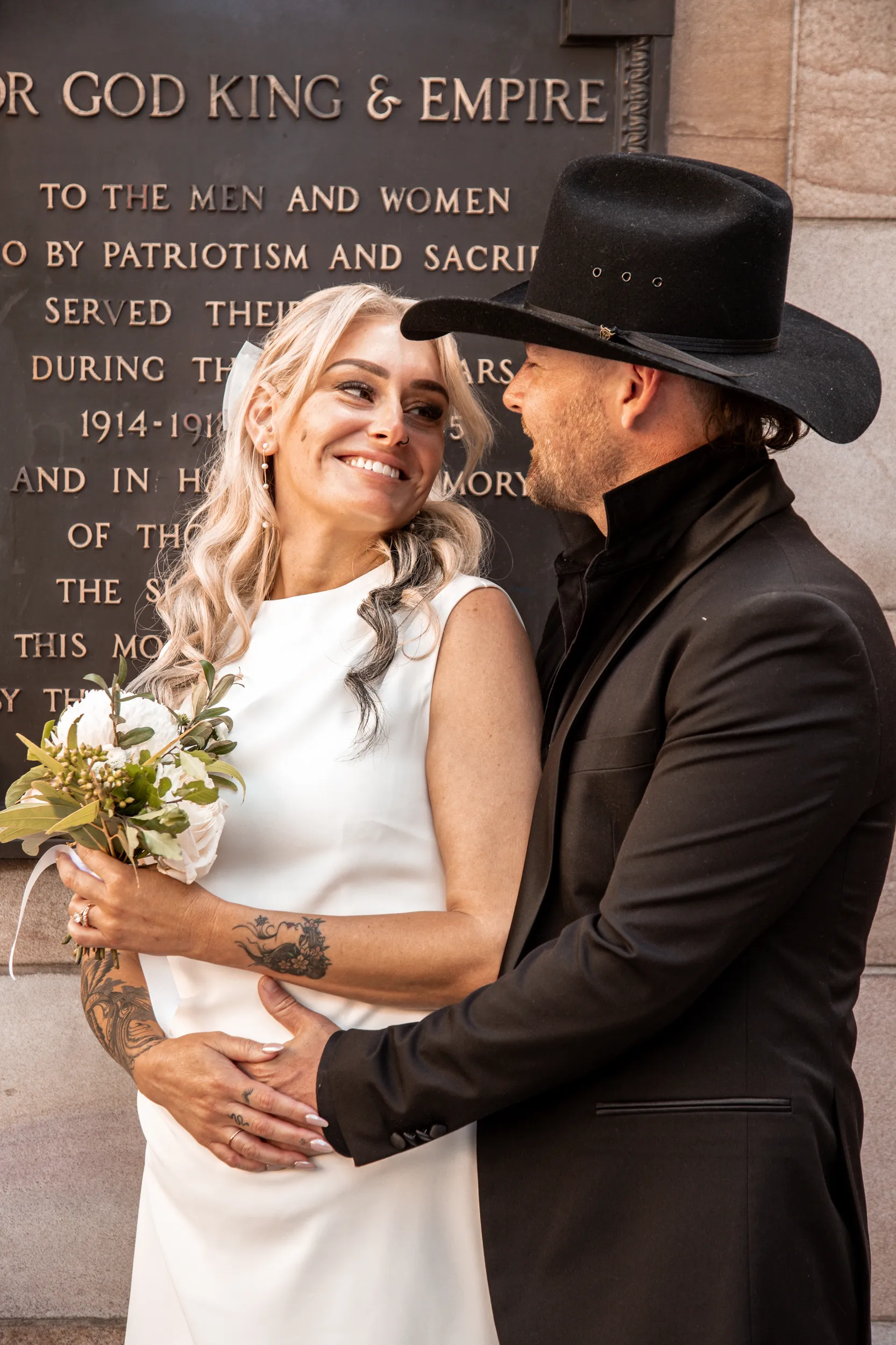Romantic wedding portrait of couple embracing in front of memorial wall with groom in cowboy hat