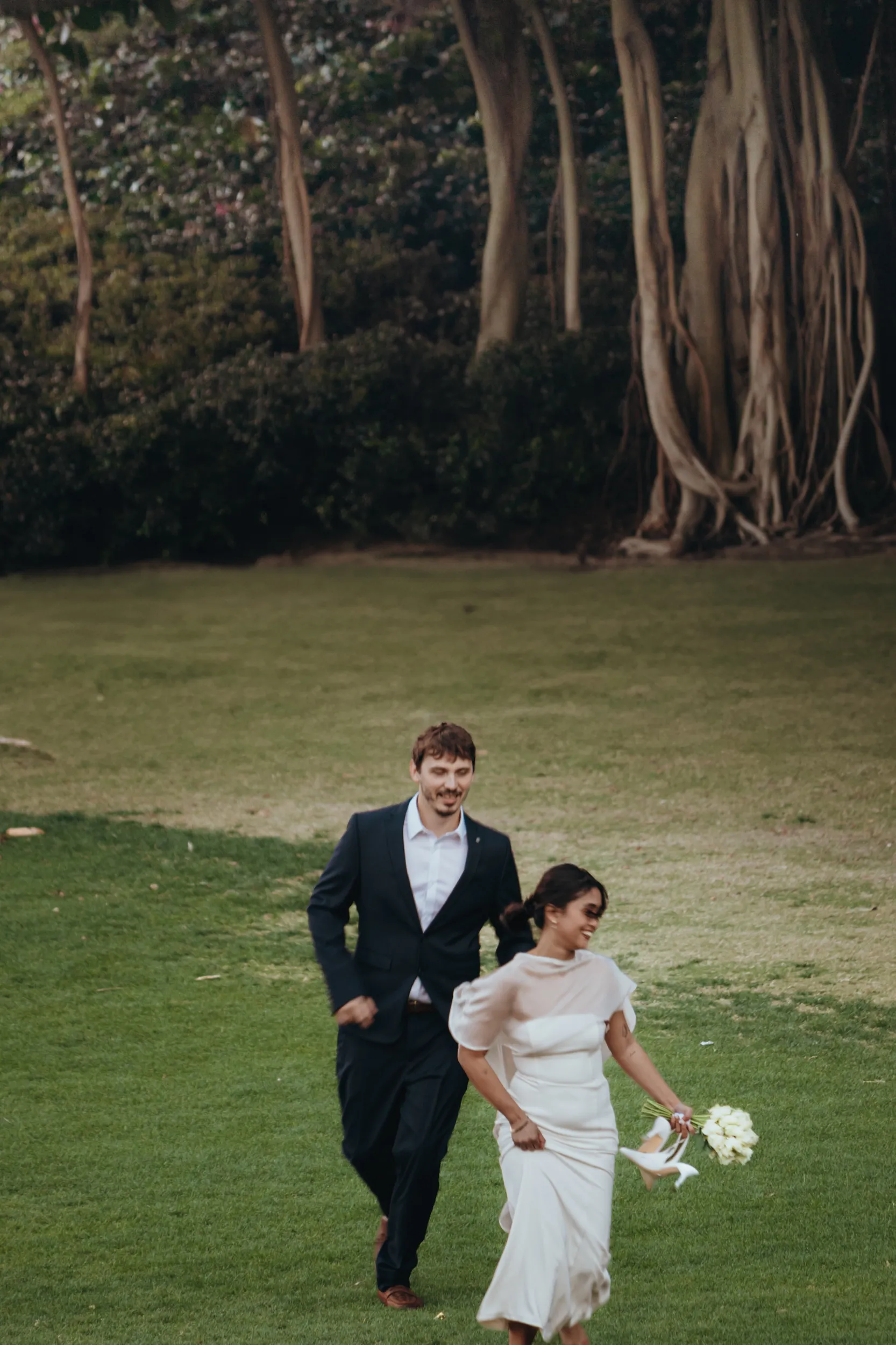 Joyful wedding couple running across grassy field during golden hour with bride in flowing white dress